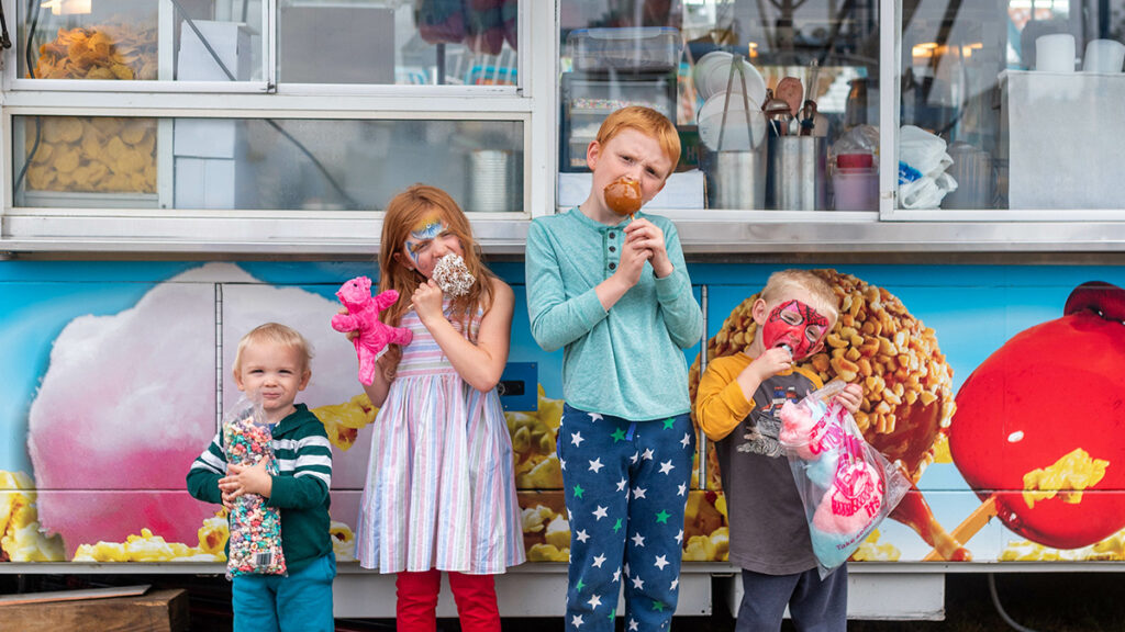 Four kids eating caramel apples and cotton candy while standing in front of a food truck.